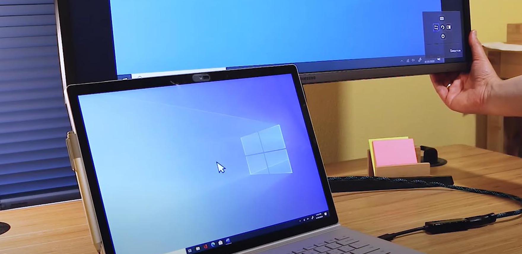 Laptop screen open under desktop monitor with a hand adjusting setting on the latter