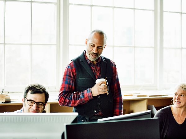 Man standing with a cup of coffee speaks with a man and a woman working on desktop computers in an office