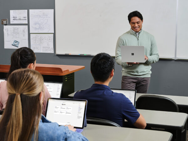 Teacher holding a laptop computer in front of a classroom of students with laptops.