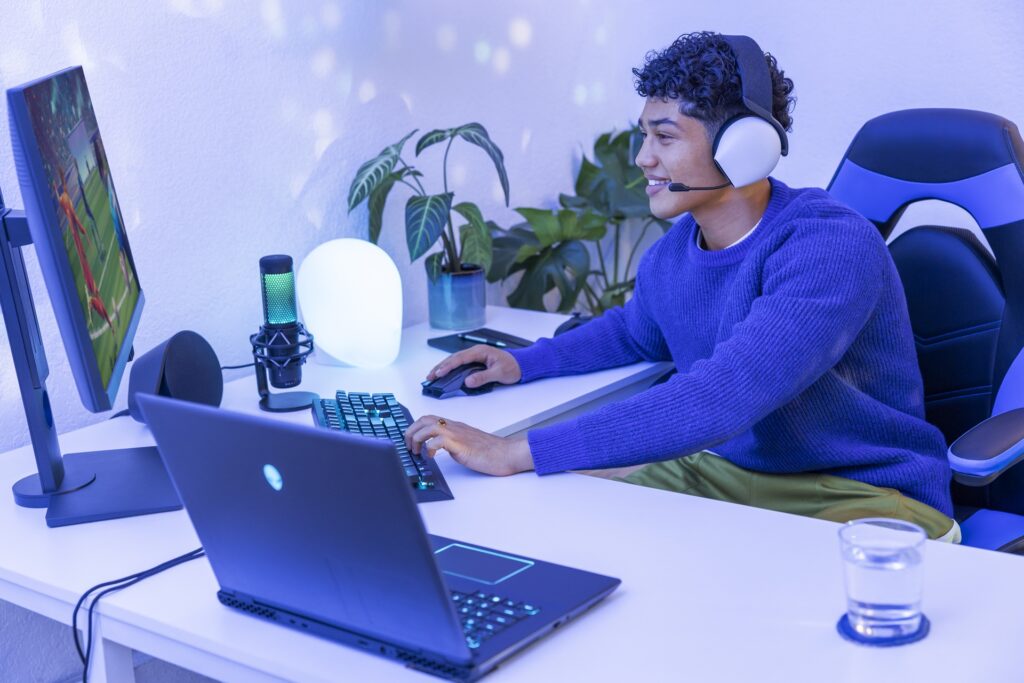 Man talking to someone over his headset while working at a desk with a laptop computer, monitor and keyboard.